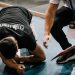 An athlete being assisted by a coach during an indoor training session on a blue floor.